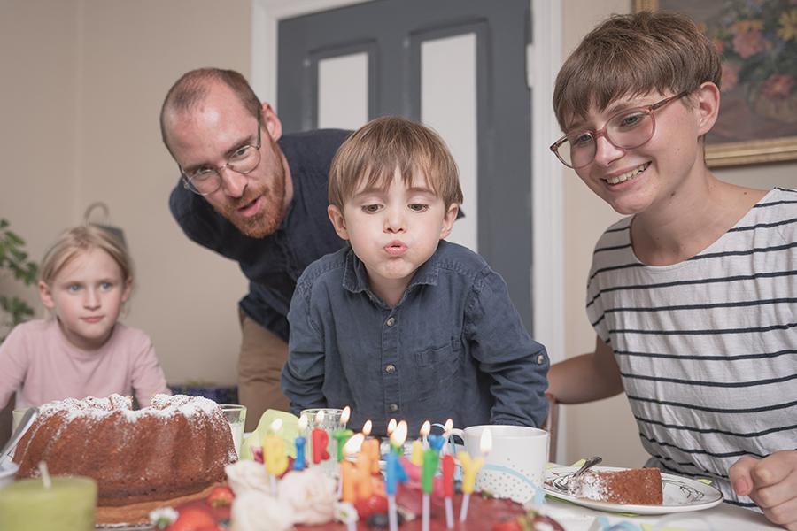 Eltern und Kinder, ein Junge und ein Mädchen sitzen gemeinsam am Tisch und feiern Geburtstag. Der Junge bläst die Kerzen auf dem Geburtstagskuchen aus, der Vater sieht ihm dabei über die Schulter. Eltern und Kinder, ein Junge und ein Mädchen sitzen gemeinsam am Tisch und feiern Geburtstag. Der Junge bläst die Kerzen auf dem Geburtstagskuchen aus, der Vater sieht ihm dabei über die Schulter.