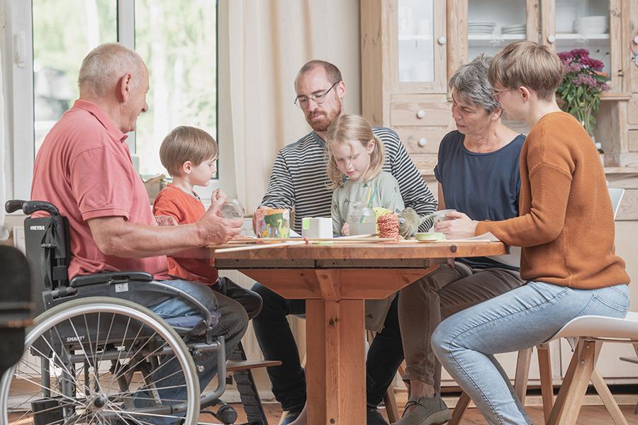 An einem runden Holztisch sitzen Großeltern, Eltern, ein Junge und ein Mädchen. Die Kinder malen und basteln. Die Erwachsenen unterhalten sich. An einem runden Holztisch sitzen Großeltern, Eltern, ein Junge und ein Mädchen. Die Kinder malen und basteln. Die Erwachsenen unterhalten sich.