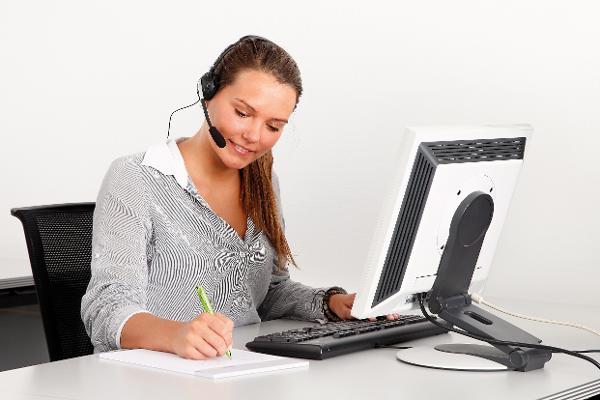 Eine junge Frau sitzt vor einem PC; sie trägt ein Headset und notiert etwas auf einem Block.