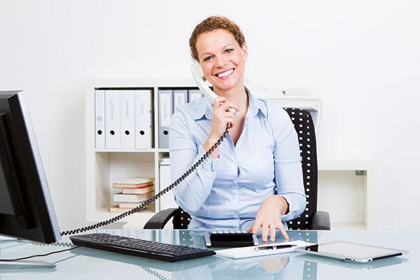 Eine junge Frau sitzt, einen Telefonhörer ans Ohr haltend, am Schreibisch und lächelt in die Kamera.
