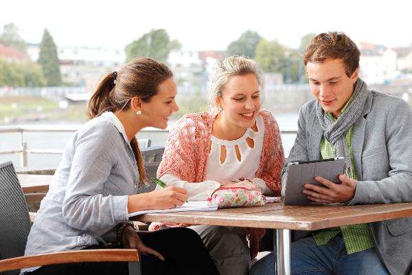 Drei junge Menschen sitzen im Freien an einem Tisch, einer hält ein Tablet, eine andere macht sich Notizen.