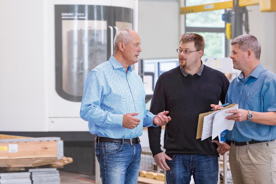 Drei Männer stehen in einer Fertigungshalle und unterhalten sich. Drei Männer stehen in einer Fertigungshalle und unterhalten sich.