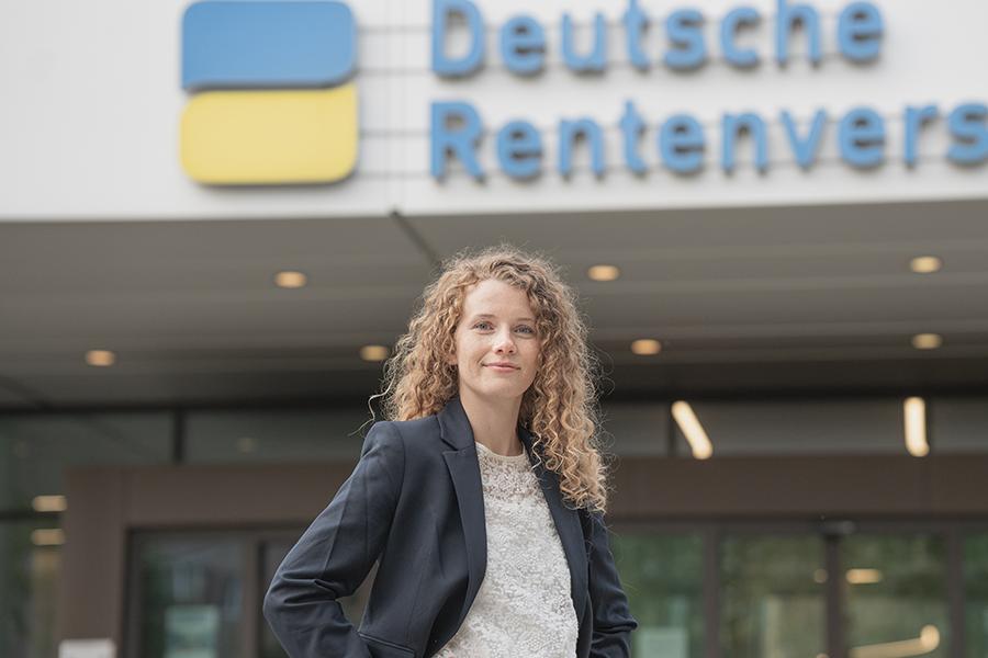 Eine junge Frau mit hellen lockigen Haaren steht vor einem Gebäude der Deutschen Rentenversicherung.