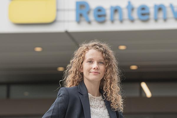 Eine junge Frau mit hellen lockigen Haaren steht vor einem Gebäude der Deutschen Rentenversicherung.  Eine junge Frau mit hellen lockigen Haaren steht vor einem Gebäude der Deutschen Rentenversicherung.