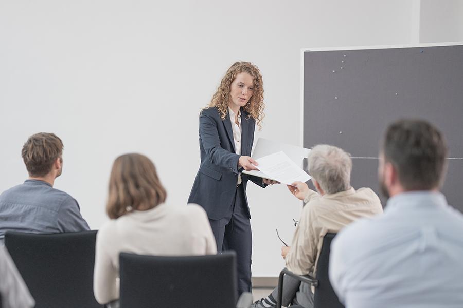 Eine junge Frau mit hellen gelockten Haaren in einem dunklen Blazer hält einen Vortrag vor Publikum. Sie händigt einem Teilnehmer Unterlagen aus. Eine junge Frau mit hellen gelockten Haaren in einem dunklen Blazer hält einen Vortrag vor Publikum. Sie händigt einem Teilnehmer Unterlagen aus.