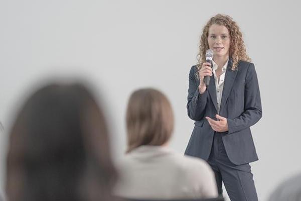 Eine junge Frau hält einen Vortrag vor Publikum. Sie hält ein Mikrofon in der Hand. Die Frau hat lockiges, helles Haar und trägt einen Blazer. Eine junge Frau hält einen Vortrag vor Publikum. Sie hält ein Mikrofon in der Hand. Die Frau hat lockiges, helles Haar und trägt einen Blazer.