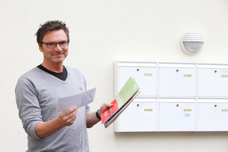 Ein Mann steht neben Briefkästen und hält eine Broschüre sowie einen Brief in der Hand.