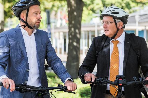 Zwei Männer im Anzug stehen mit ihren Fahrrädern an einem Weg und unterhalten sich. Sie tragen beide einen Fahrradhelm. Zwei Männer im Anzug stehen mit ihren Fahrrädern an einem Weg und unterhalten sich. Sie tragen beide einen Fahrradhelm.