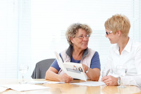 Eine ältere Frau hält eine Rentenberatungsbroschüre, neben ihr sitzt eine Beraterin. Eine ältere Frau hält eine Rentenberatungsbroschüre, neben ihr sitzt eine Beraterin.