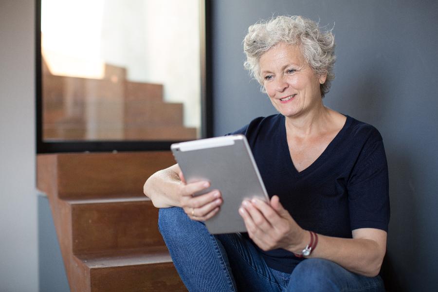 Eine lächelnde Seniorin sitzt auf der Treppe ihres Haueses und bedient ein Tablet