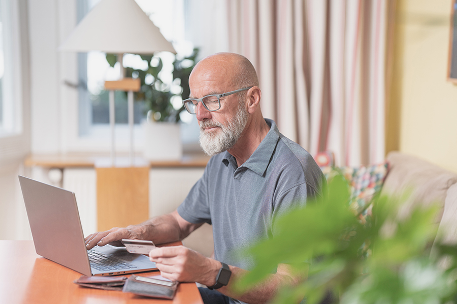 Älterer Herr mit Brille und Bart sitzt vor dem Laptop und hat seine EC-Karte in der Hand Älterer Herr mit Brille und Bart sitzt vor dem Laptop und hat seine EC-Karte in der Hand