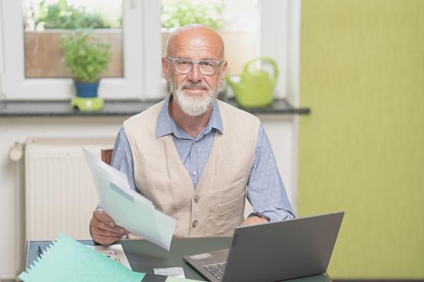 Älterer Herr sitzt mit Unterlagen vor seinem Laptop am Küchentisch Älterer Herr sitzt mit Unterlagen vor seinem Laptop am Küchentisch