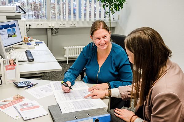 In einem Büro sitzen zwei Frauen an einem Tisch. Die eine Frau berät die andere und lächelt sie an. In einem Büro sitzen zwei Frauen an einem Tisch. Die eine Frau berät die andere und lächelt sie an.