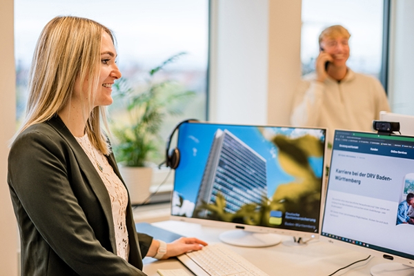 Das Bild zeigt eine Frau, die auf zwei Bildschirme blickt. Dahinter steht ein telefonierender Mann. Das Bild zeigt eine Frau, die auf zwei Bildschirme blickt. Dahinter steht ein telefonierender Mann.