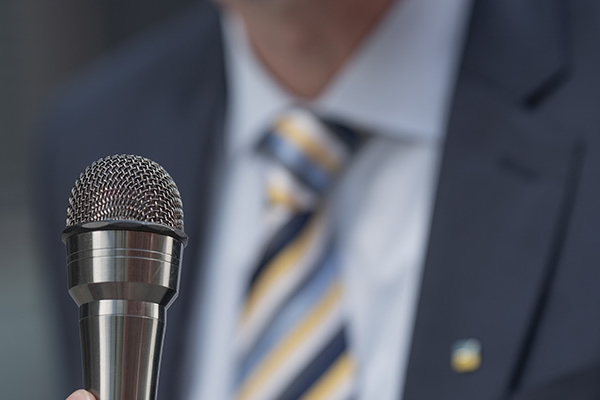 Hier ist ein Bild vonn einem Mikrofon zu sehen, im Hintergrund steht eine Person die enen Schlips trägt Hier ist ein Bild vonn einem Mikrofon zu sehen, im Hintergrund steht eine Person die enen Schlips trägt