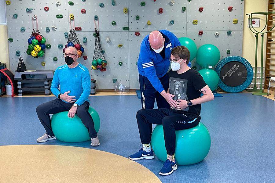 Ein Therapeut leitet zwei auf Gymnastikbällen sitzende Rehabilitanden zu Atemübungen an. Ein Therapeut leitet zwei auf Gymnastikbällen sitzende Rehabilitanden zu Atemübungen an.