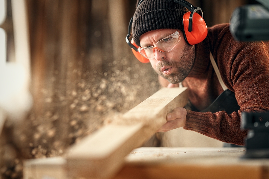 Das Bild zeigt einen Mann mit brauner Mütze und leuchtend rotem Gehörschutz. Er blickt nach vorne, sägt einen Holzbalken und pustet die Sägespäne gleichzeitig weg.