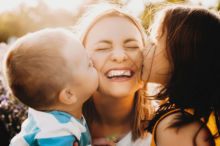 Das Bild zeigt eine Frau, die von zwei Seiten zugleich von zwei Kindern ins Gesicht geküsst wird. Von einem kleinem Jungen von links und einem etwas älterem Mädchen von rechts. Die Frau lacht dabei.