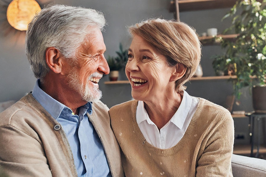 Das Bild zeigt eine älteres Paar in häuslicher Umgebung, das sich gegenseitig anlacht. Beide tragen mit beiger Oberbekleidung. Der Mann (links) hat graue Haare, einen Bart und ein blaues Hemd, die Frau (rechts) blondgraue Haare und ein weißes Hemd. Das Bild zeigt eine älteres Paar in häuslicher Umgebung, das sich gegenseitig anlacht. Beide tragen mit beiger Oberbekleidung. Der Mann (links) hat graue Haare, einen Bart und ein blaues Hemd, die Frau (rechts) blondgraue Haare und ein weißes Hemd.
