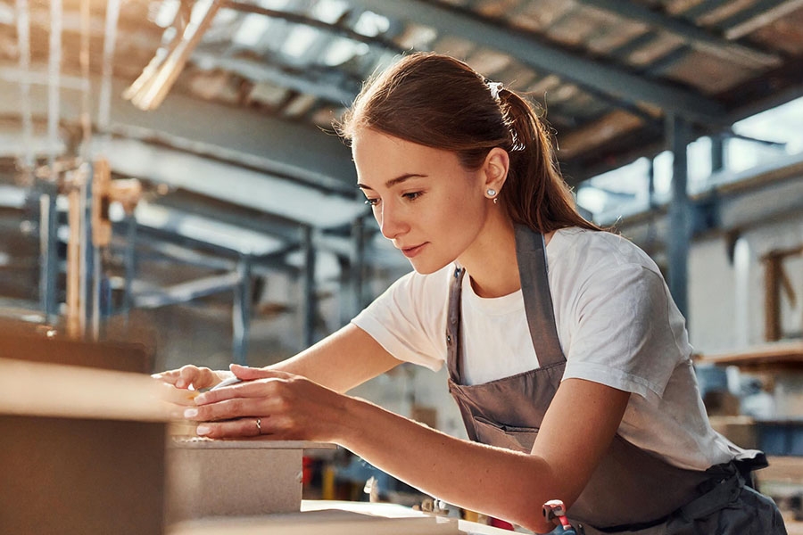 Das Bild zeigt eine Frau mit weißem Shirt und grauer Arbeitsschürze, die in einer Fabrikhalle Kartons befüllt. Das Bild zeigt eine Frau mit weißem Shirt und grauer Arbeitsschürze, die in einer Fabrikhalle Kartons befüllt.