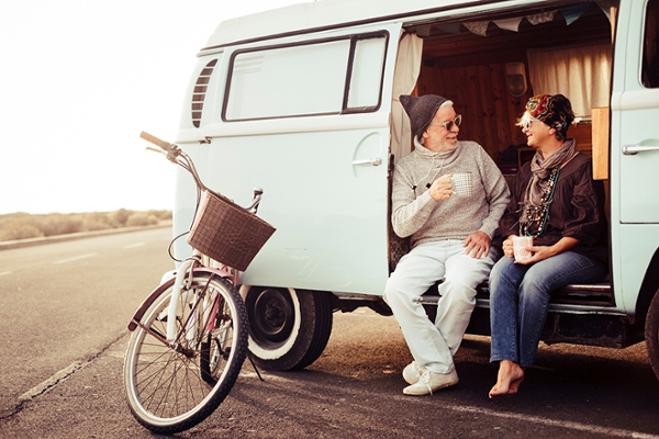 Ein Paar sitzt mit einer Kaffeetassen in der Hand vor der offenen Tür eines Campingbullis und spricht miteinander. Ein Paar sitzt mit einer Kaffeetassen in der Hand vor der offenen Tür eines Campingbullis und spricht miteinander.