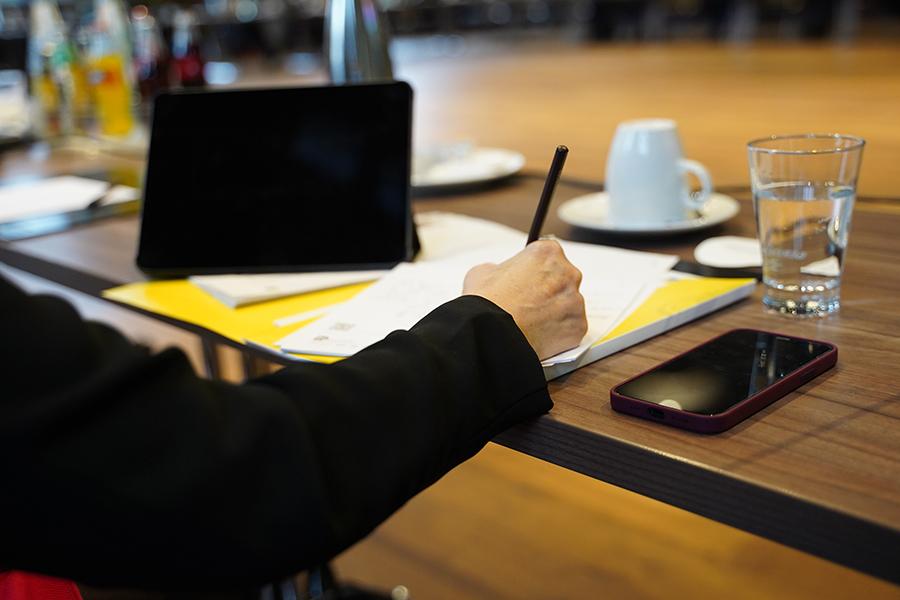 Das Bild zeigt einen Schreibvorgang am Tisch, auf dem sich auch ein Handy und ein Laptop befinden. Das Bild zeigt einen Schreibvorgang am Tisch, auf dem sich auch ein Handy und ein Laptop befinden.