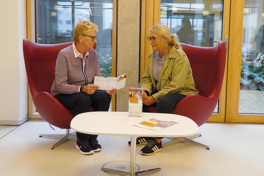 Das Bild zeigt zwei Frauen, die sich in weinroten Sesseln gegenübersitzen. Die Frau auf der linken Seite hält einen aufgeklappten Flyer in der Hand. Das Bild zeigt zwei Frauen, die sich in weinroten Sesseln gegenübersitzen. Die Frau auf der linken Seite hält einen aufgeklappten Flyer in der Hand.