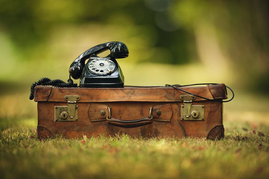 Das Bild zeigt einen alten braunen Koffer, auf dem ein schwarzes Telefon mit Wählscheibe steht. Beides zusammen steht auf einer grünen Wiese.