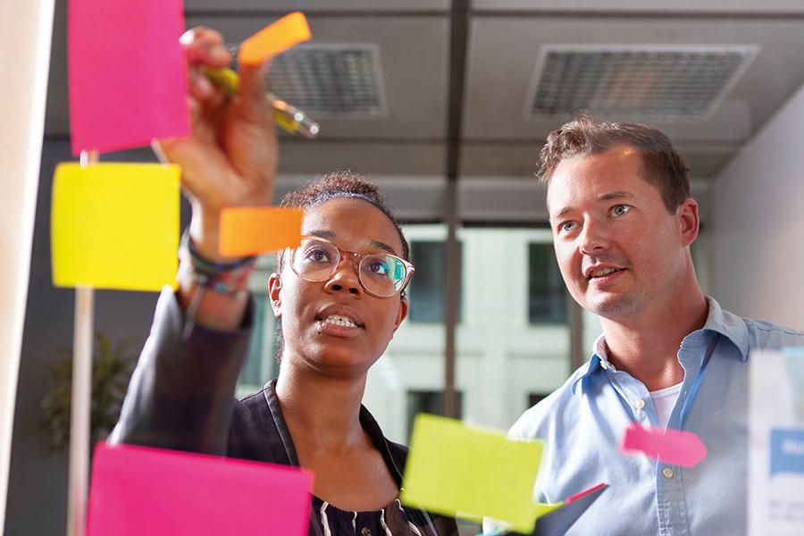 Das Bild zeigt eine Frau und einen Mann, die an einer Glaswand stehen und bunte Zettel ankleben. Das Bild zeigt eine Frau und einen Mann, die an einer Glaswand stehen und bunte Zettel ankleben.