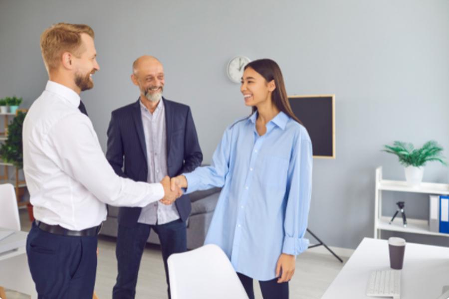Das Bild zeigt zwei Männer, die eine junge Frau per Handschlag in einem Büro begrüßen. Das Bild zeigt zwei Männer, die eine junge Frau per Handschlag in einem Büro begrüßen.