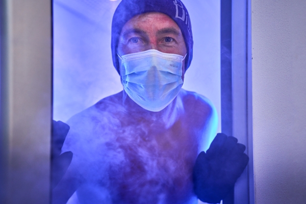 Ein Mann, oberkörperfrei, steht mit Handschuhen und Maske in der Tür zu einer Kältekammer. Er ist umgeben von blauem Licht und aufsteigendem Nebel. Ein Mann, oberkörperfrei, steht mit Handschuhen und Maske in der Tür zu einer Kältekammer. Er ist umgeben von blauem Licht und aufsteigendem Nebel.