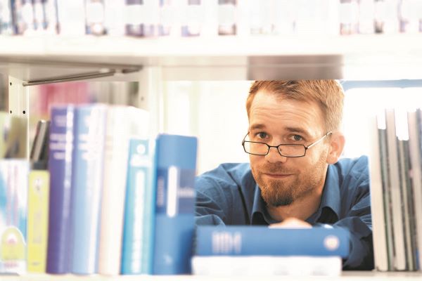 Ein Mann mit Brille schaut einen zwischen Büchern hindurch an. Ein Mann mit Brille schaut einen zwischen Büchern hindurch an.