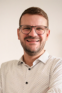 Ein Mann mit Brille, kurzen Haaren und hellem Hemd mit hochgekrempelten Ärmeln lächelt in die Kamera. Ein Mann mit Brille, kurzen Haaren und hellem Hemd mit hochgekrempelten Ärmeln lächelt in die Kamera.