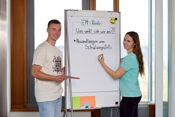 Zwei junge Leute stehen vor einem Flipchart mit dem Titel "EM-Rente". Beide lächeln in die Kamera. Zwei junge Leute stehen vor einem Flipchart mit dem Titel "EM-Rente". Beide lächeln in die Kamera.