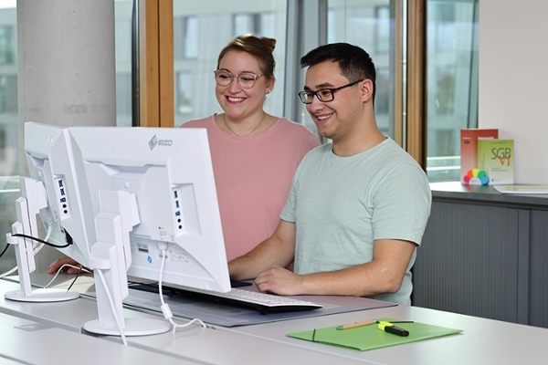 Zwei lächelnde junge Leute stehen zusammen vor zwei Computermonitoren und blicken darauf.