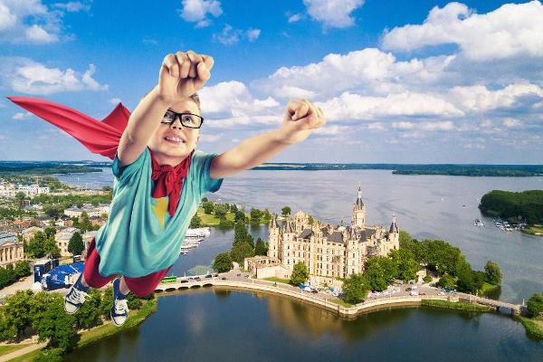 Ein Junge mit rotem Cape fliegt in Superhelden-Pose über das Schweriner Schloss und den Schweriner See.