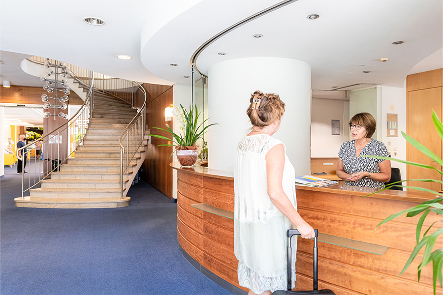 Eine Mitarbeiterin der Klinik begrüßt eine Patientin an der Rezeption. Eine Mitarbeiterin der Klinik begrüßt eine Patientin an der Rezeption.