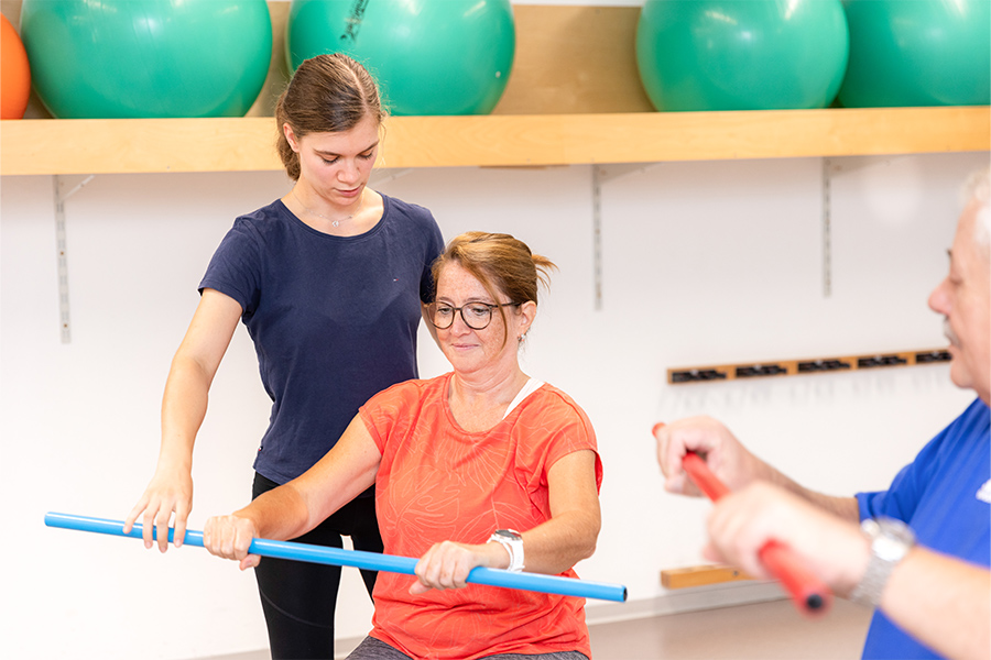Eine Therapeutin korrigiert die Haltung einer Patientin beim Training mit dem Turnstab. Eine Therapeutin korrigiert die Haltung einer Patientin beim Training mit dem Turnstab.