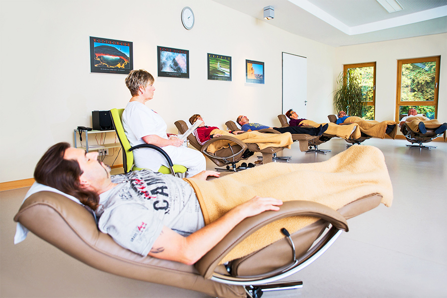 Mehrere Patienten entspannen auf Liegesesseln, während eine Therapeutin etwas vorliest. Mehrere Patienten entspannen auf Liegesesseln, während eine Therapeutin etwas vorliest.