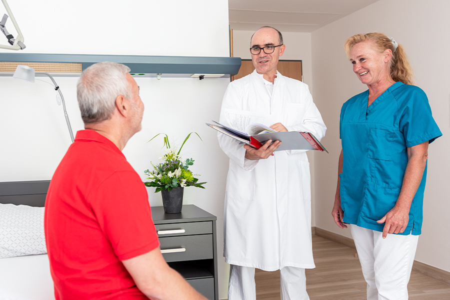 Ein Patient sitzt in seinem Zimmer auf seinem Bett und unterhält sich mit einem Arzt und einer Frau vom Pflegepersonal, die ihm gegenüber stehen. Ein Patient sitzt in seinem Zimmer auf seinem Bett und unterhält sich mit einem Arzt und einer Frau vom Pflegepersonal, die ihm gegenüber stehen.
