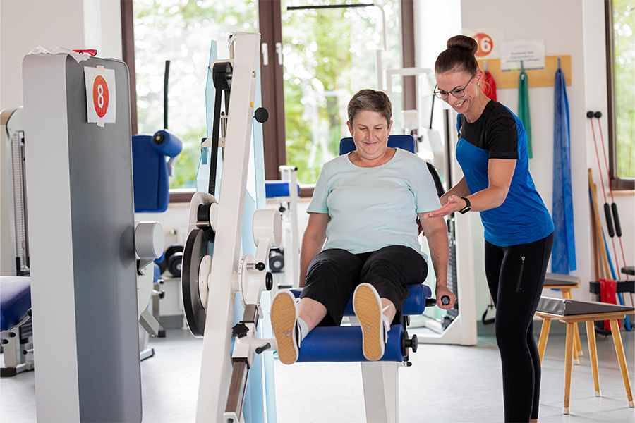 Eine Therapeutin unterstützt eine Patientin beim Training an einem Trainingsgerät für die Beine. Eine Therapeutin unterstützt eine Patientin beim Training an einem Trainingsgerät für die Beine.