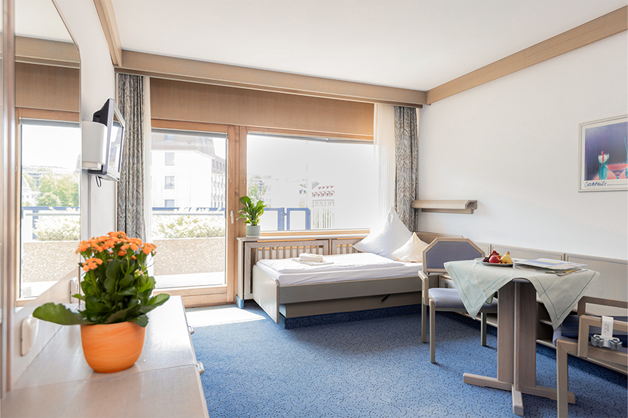 Ein beispielhaftes Patientenzimmer mit großen Fenstern, Balkon, Fernseher, Bett und Sitzecke. Ein beispielhaftes Patientenzimmer mit großen Fenstern, Balkon, Fernseher, Bett und Sitzecke.