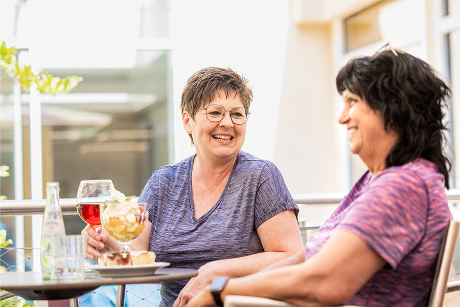 Zwei Patientinnen sitzen draußen an einem Tisch mit Getränken, Obstsalat und Kuchen und unterhalten sich. Zwei Patientinnen sitzen draußen an einem Tisch mit Getränken, Obstsalat und Kuchen und unterhalten sich.