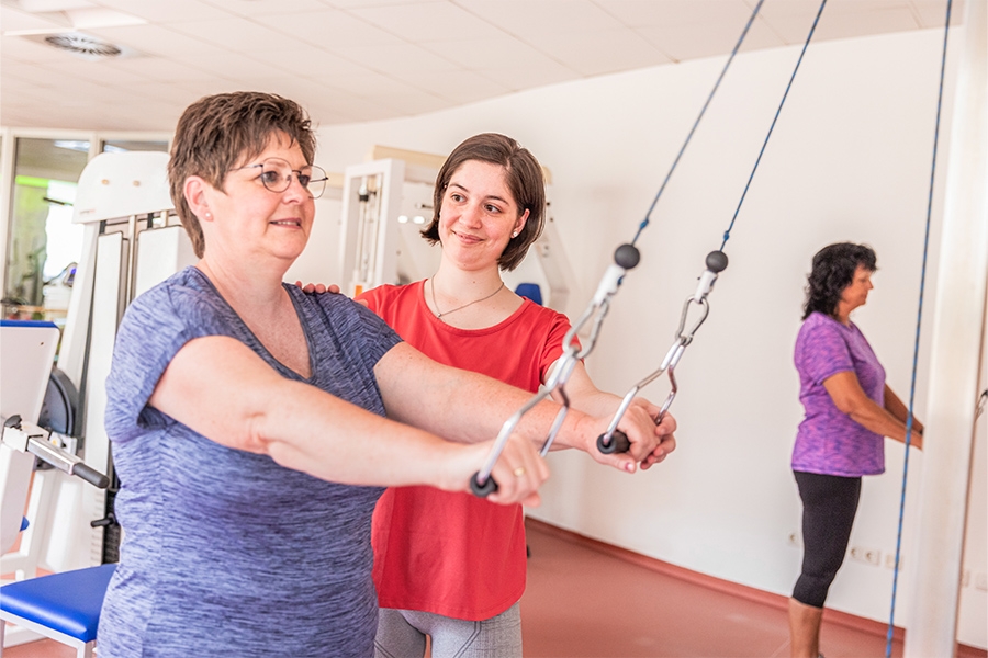 Eine Therapeutin korrigiert die Haltung einer Patientin beim Training an einem Trainingsgerät. Eine Therapeutin korrigiert die Haltung einer Patientin beim Training an einem Trainingsgerät.
