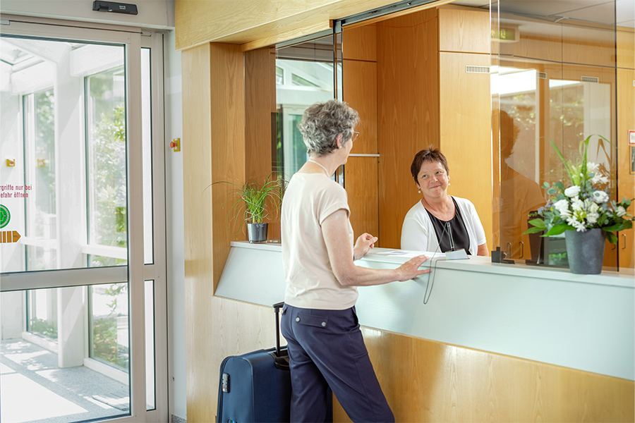Eine Patientin mit Koffer wird von einer Mitarbeiterin der Klinik am Empfang begrüßt. Eine Patientin mit Koffer wird von einer Mitarbeiterin der Klinik am Empfang begrüßt.