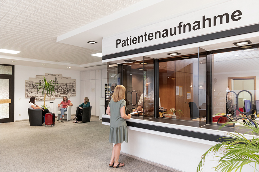 Eine Frau spricht mit einem Mitarbeiter an der Patientenaufnahme. Im Hintergrund sitzen weitere Patientinnen im Wartebereich. Eine Frau spricht mit einem Mitarbeiter an der Patientenaufnahme. Im Hintergrund sitzen weitere Patientinnen im Wartebereich.