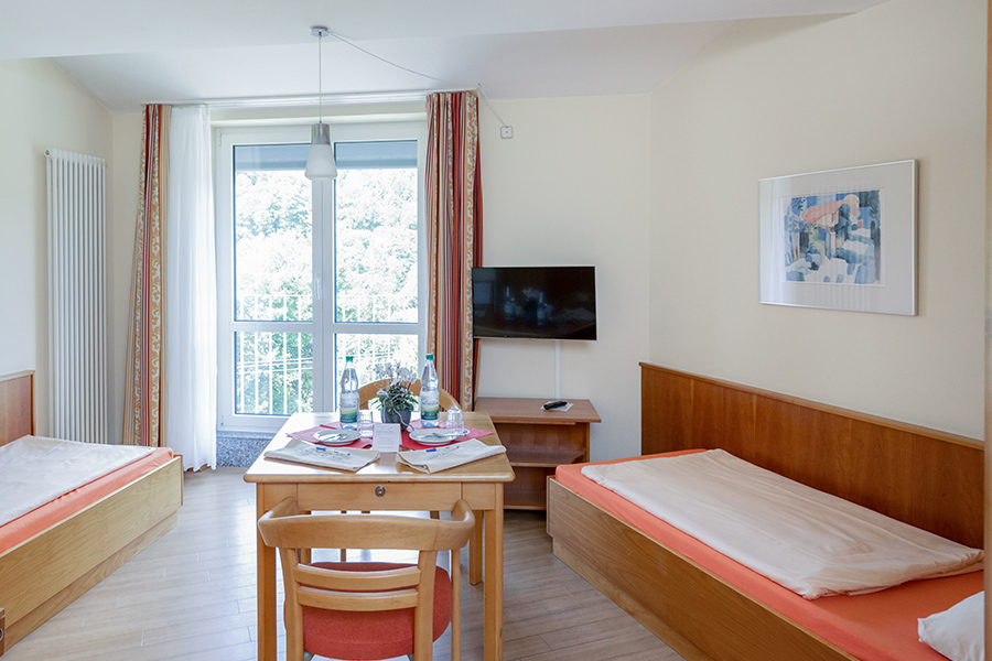 Ein beispielhaftes Patientenzimmer mit zwei Betten, Fernseher und Sitzbereich mit einem gedeckten Tisch. Ein beispielhaftes Patientenzimmer mit zwei Betten, Fernseher und Sitzbereich mit einem gedeckten Tisch.