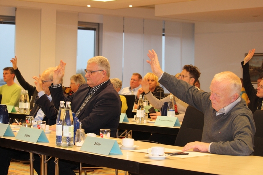 Mehrere Personen sitzen neben- und hintereinander an Tischen und heben ihre Hand zur Abstimmung. Mehrere Personen sitzen neben- und hintereinander an Tischen und heben ihre Hand zur Abstimmung.