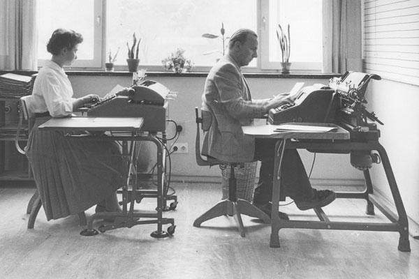 Das schwarz-weiß Foto zeigt eine Frau und einen Mann im Kleidungsstil der 50er Jahre, die an zwei Tischen hintereinander vor jeweils einer Schreibmaschine sitzen.