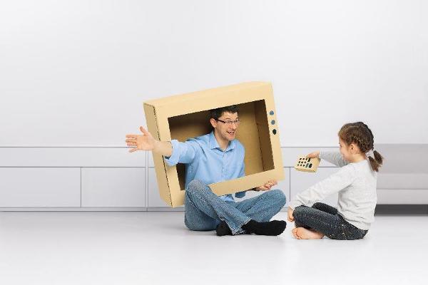 Vater sitzt in einem Fernseher aus Karton und ein Mädchen hält eine selbstgebastelte Fernbedienung in seine Richtung Vater sitzt in einem Fernseher aus Karton und ein Mädchen hält eine selbstgebastelte Fernbedienung in seine Richtung
